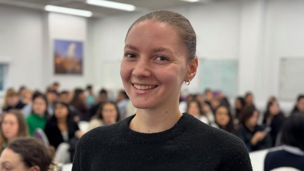 Chloe Wilson smiles at the camera. She's wearing a black top and hooped earrings. She's standing in front of students who are blurred. They're sitting at tables in a white room with some pictures on the wall. 