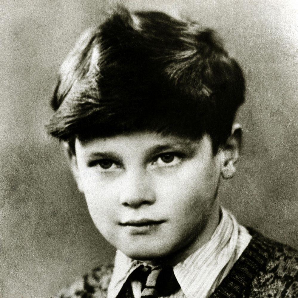 A black and white image of Sir Roger Moore as a child. He has a serious but pensive expression on his face and is wearing a stripy shirt, a tie and a patterned knitted jumper. He has a full head of dark hair.