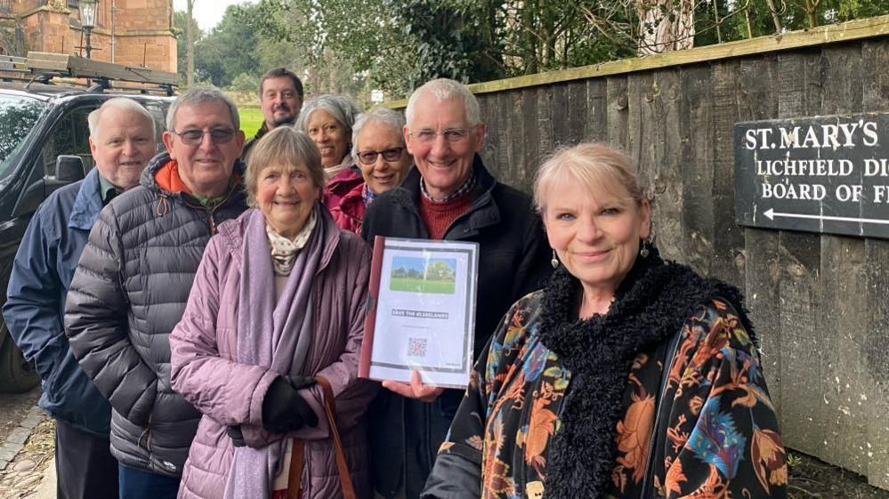 Photo of the seven campaigners stood next to a sign reading 'St Mary's Lichfield'. They are all wearing winter coats, and Mark Underwood, a man with white hair and glasses, is holding the petition booklet to be delivered to the diocese. They are standing on a street with a wooden fence running along the right hand side of the image. 