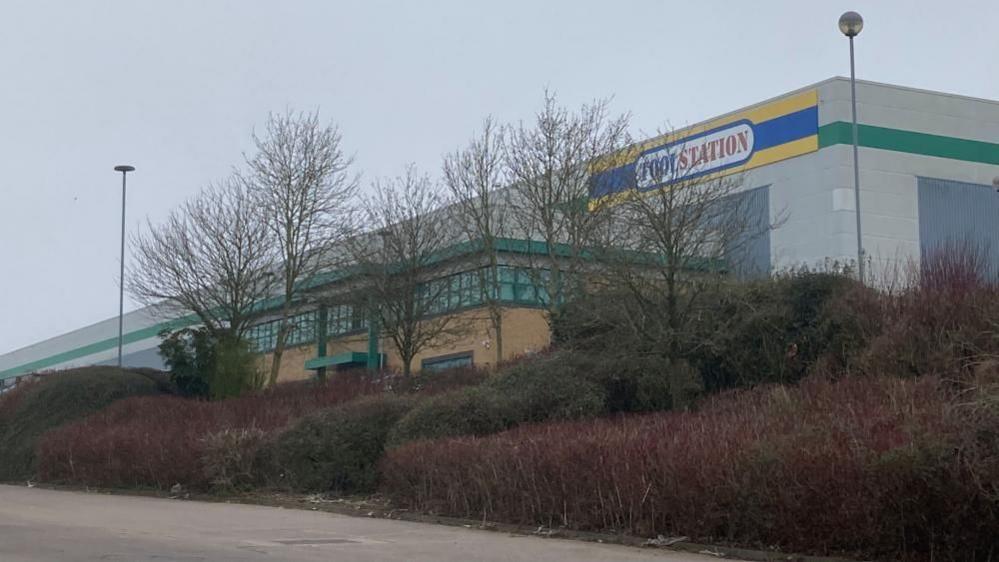 Large grey warehouse building with single-storey entrance area and "Toolstation" sign. There are trees and bushes outside.