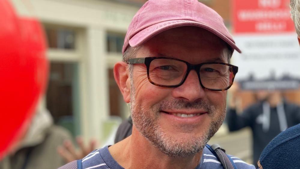 Graham Mellor with very short grey hair and beard, wearing glasses and a pink hat. Other campaigners are visible behind him.