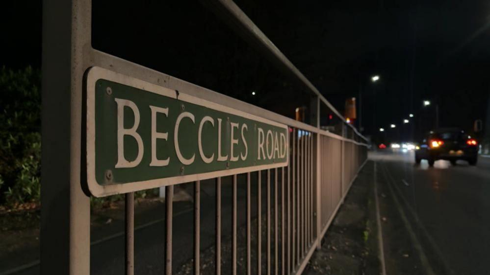 A road sign stating Beccles Road, lit by car headlights. The sign shows white text and border on a green background and the sign is affixed to metal railings. Car tail lamps and headlights can be seen to the right of the image.