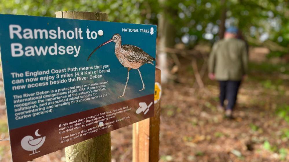 There's a signpost on a wooden fence which says Ramsholt to Bawdsey - the England Coast path - you can now enjoy 3 miles (4.8km) of brand new access beside the river Deben, It has a picture then of a Stone Curlew on it the side. In the distance to the right f the sign you can see tow blurred walkers.