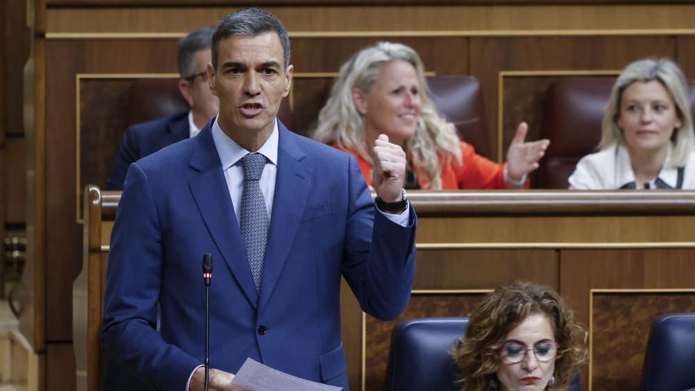 A man in a blue suit speaks in the Spanish parliament raising his left hand
