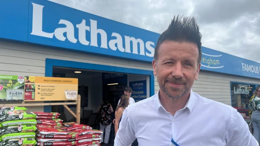 John Goldie, wearing a white shirt, standing outside Lathams of Potter Heigham. Next to him is a stack of bagged compost, and the signage of the shop. John has a close cropped beard and moustache, and has spiked hair.