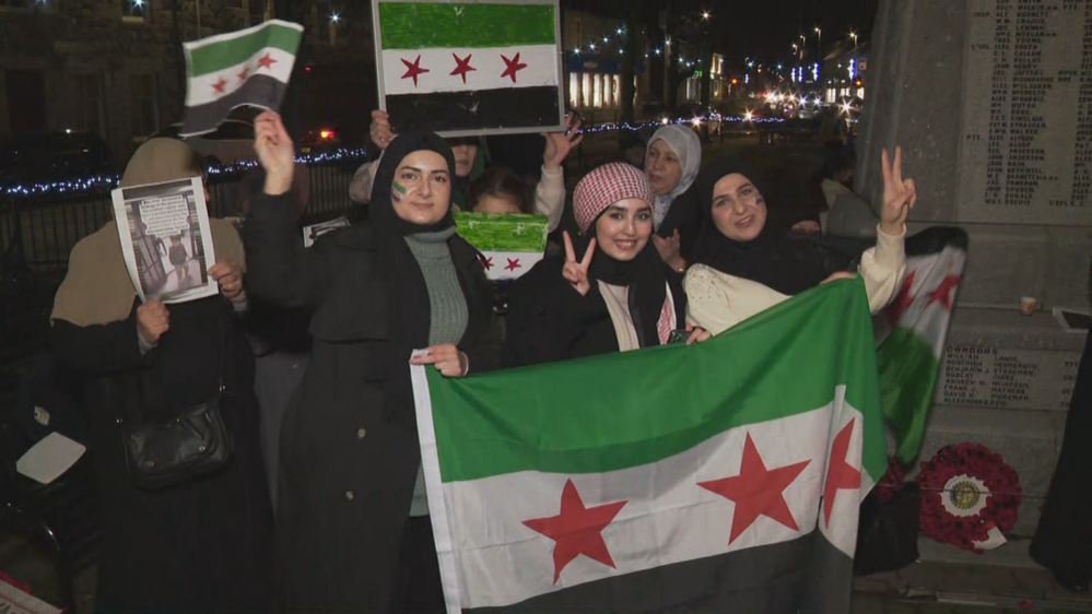 People celebrate with Syrian flags in Inverurie. It's dark and they are crowded in front of a monument showing peace signs to the camera.