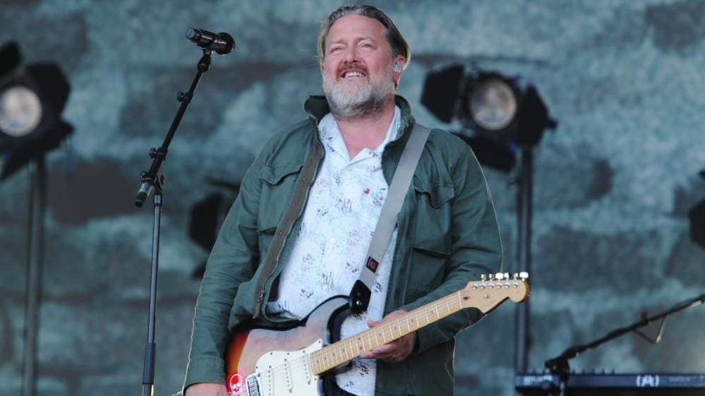 Guy Garvey is smiling on the Pyramid stage at Glastonbury. He has grey and brown hair and a grey beard and is wearing a white shirt with a floral pattern and a dark blue-green denim utility jacket. He is holding a guitar and some lighting can be seen behind him.