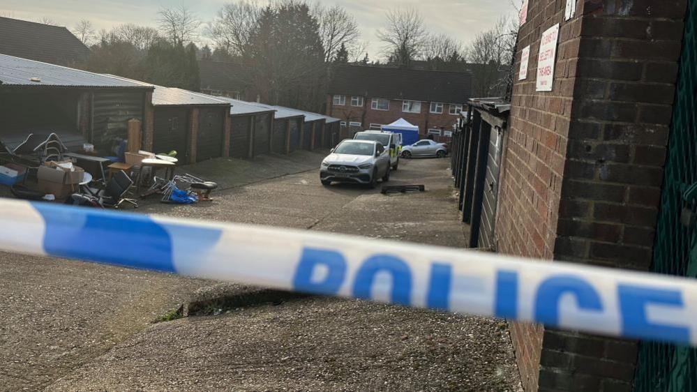 Police tape in from of a row of garages, police vehicles and a forensic tent. A row of houses are at the end of the row. 