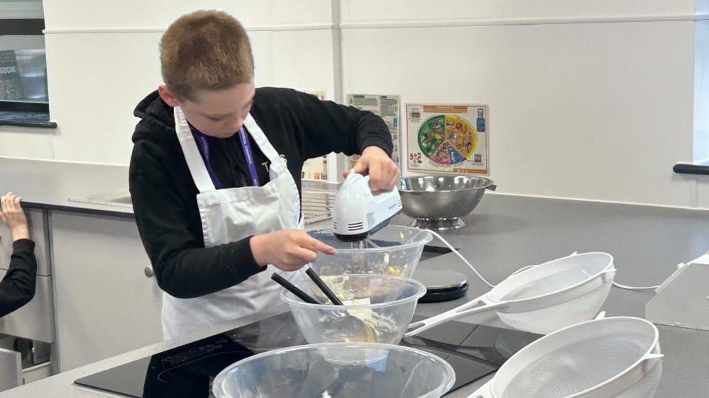 Tyler, using a whisk, in a school kitchen, with a number of clear bowls in front of him, he is looking down. There are two sieves on the counter and a silver bowl behind him. He is wearing a black jumper, with a white apron over the top. 