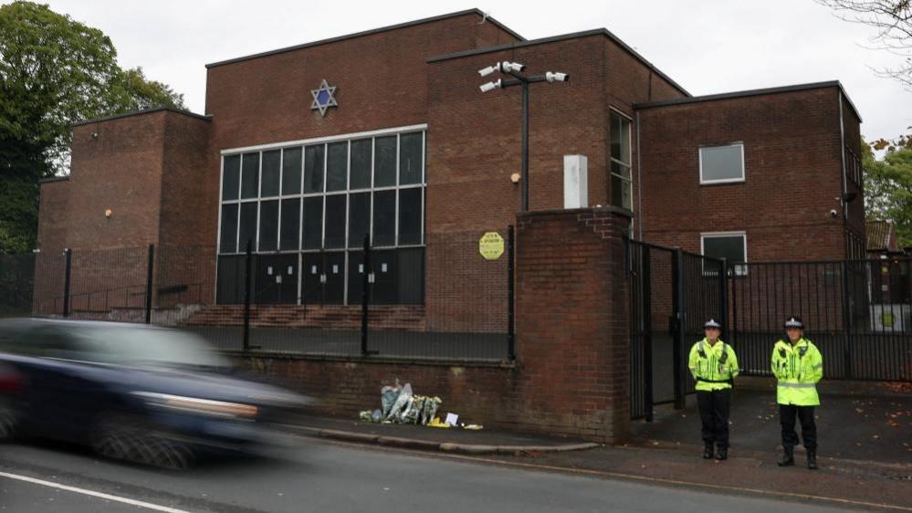 Two police officers in high-vis yellow jackets stand with their hands behind their backs in front of a dark brick building with a Star of David on the front of it. 