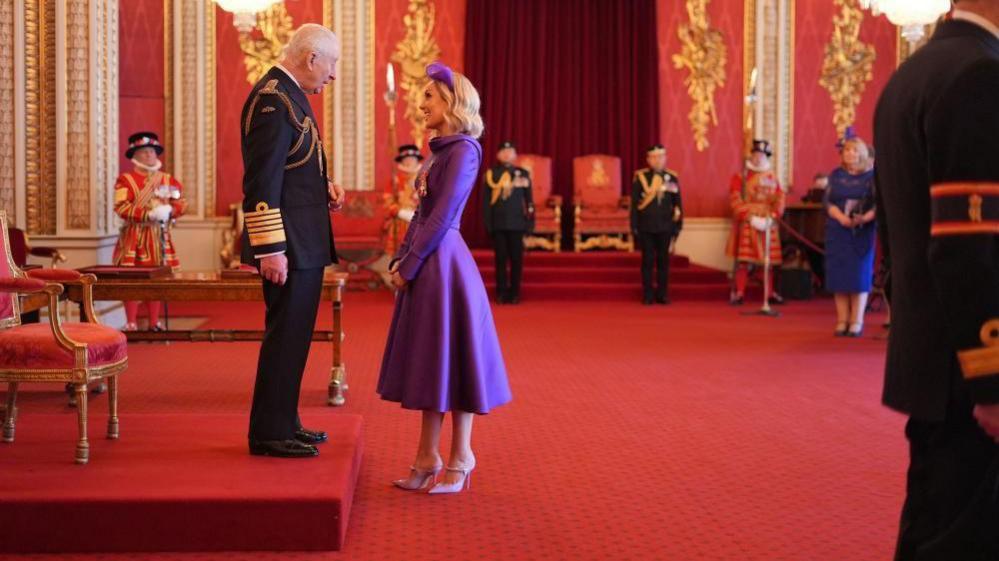 Amy Dowden stands in front of King Charles during the investiture ceremony at Buckingham Palace. He is standing on a red plinth and she is smiling and talking to him as she stands on a red carpet. He is in his naval uniform. There are a number of people in the background, including several Beefeaters.