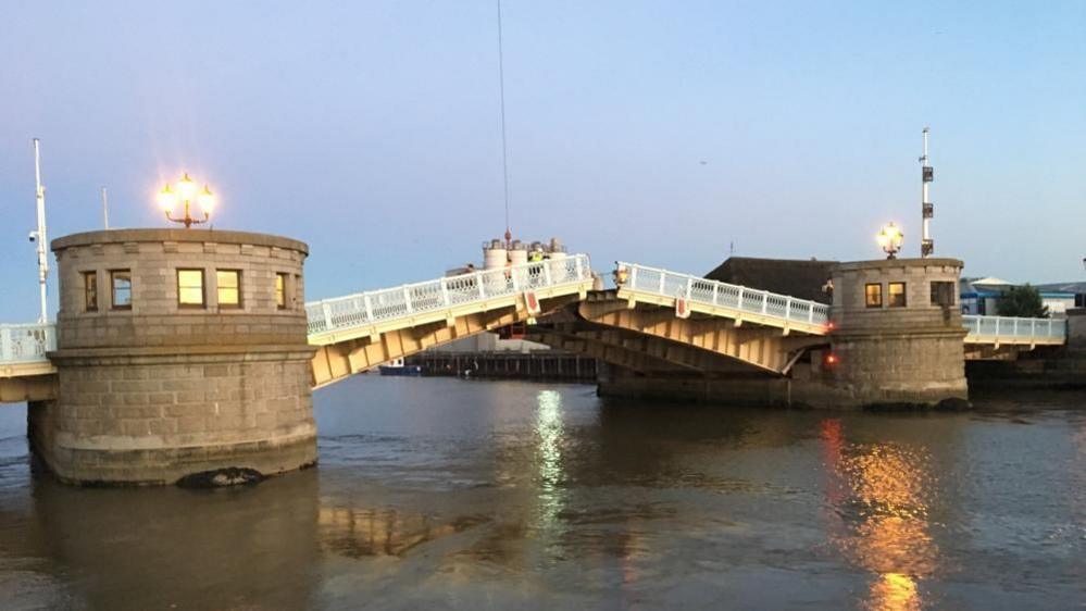 Haven Bridge, viewed from the northwest, with the bridge piers lit and the spans partially raised. Behind the west-most bridge pier is The Ice House and some flour silos at the Semolina Mill.