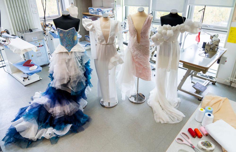 Four dress designs are tacked onto mannequins in a work shop. The first one has a blue sparkly bodice with a blue and white mesh skirt. The second is white and has a deep V neck and asymmetric detailing in the shoulders and skirt. The second  is light pink and about knee length with a very deep V down to about the hips. The fourth has white balls about the size of a fist going diagonally across the dress. It has a short skirt with a longer mesh skirt over the top.