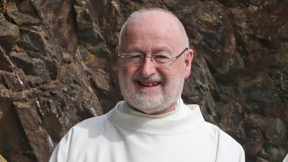 Fr John Murray. He is smiling at the camera. Fr Murray is bald and is wearing glasses. He has a white beard and is wearing white robes. Behind him is a rockface.