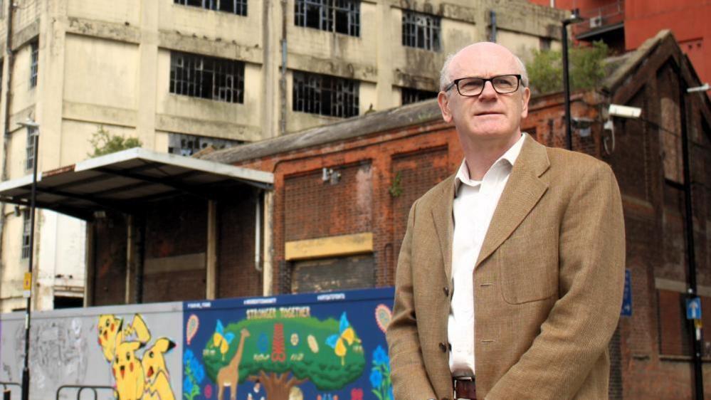 Neil McDonald wearing a brown jacket and glasses. He is standing in front of some graffiti on a derelict building and looking off in the distance. 