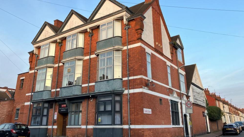 Three storey office block with red brick, white, grey and black windows. A 20 mph sign is to the right of the building