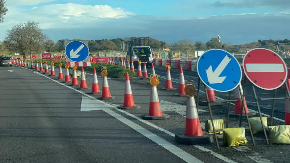 The picture shows a section of the A12 undergoing road works. The roads are lined with numerous orange-and-white traffic cones, many of which have small yellow warning lights attached. Two large signs showing drivers should go one way and that other sign is no entry.