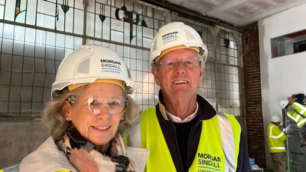 Bruce and Libby Sturrock wearing high visibility jackets, hard hats and safety glasses, standing in the former menswear department with historic stained-glass windows, bearing the initial P for Palmers, in the background.