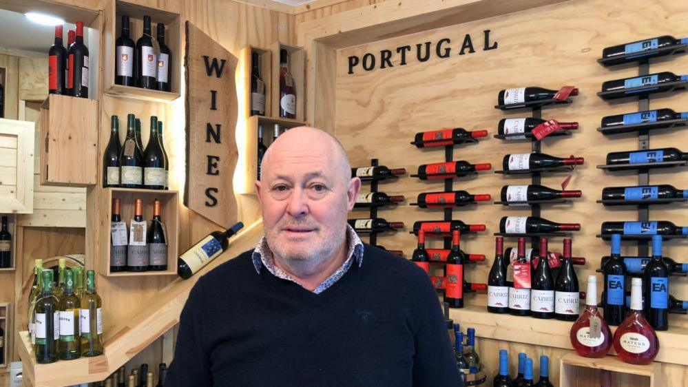 A man is standing in front of display of Portuguese and Spanish wines. He is wearing a blue jumper with a shirt underneath. 