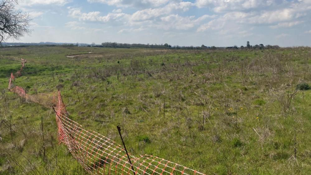 Large farm field with red plastic fencing in the foreground held up by thin metal posts. Trees are visible in the background.