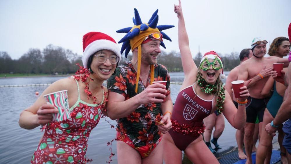 Three people, two women with a man in the centre, wearing fancy dress hats and swimming gear while smiling for the camera and clutching drinks