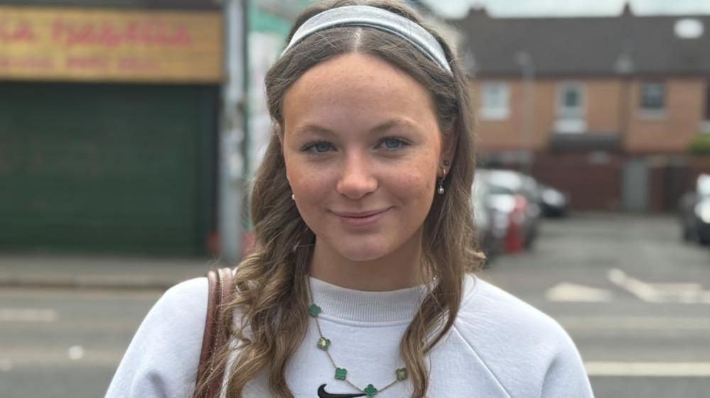 Naoise McCrory is wearing a grey headband and has her long, light coloured hair curled. She is smiling at the camera. On her right shoulder she is holding a brown leather bag strap and is wearing a grey crewneck jumper with a black Nike tick in the centre. She is stood in front of a closed shop front and an alleyway but the background is blurred.