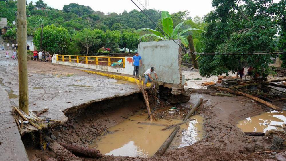 Men stand near a damaged bridge in Jalcocotan, Nayarit state. Part of the bridge has collapsed and pipes and tree trunks can be seen in a big, water-filled hole where the bridge would have ended. Residents look on as a man pulls debris from the hole.