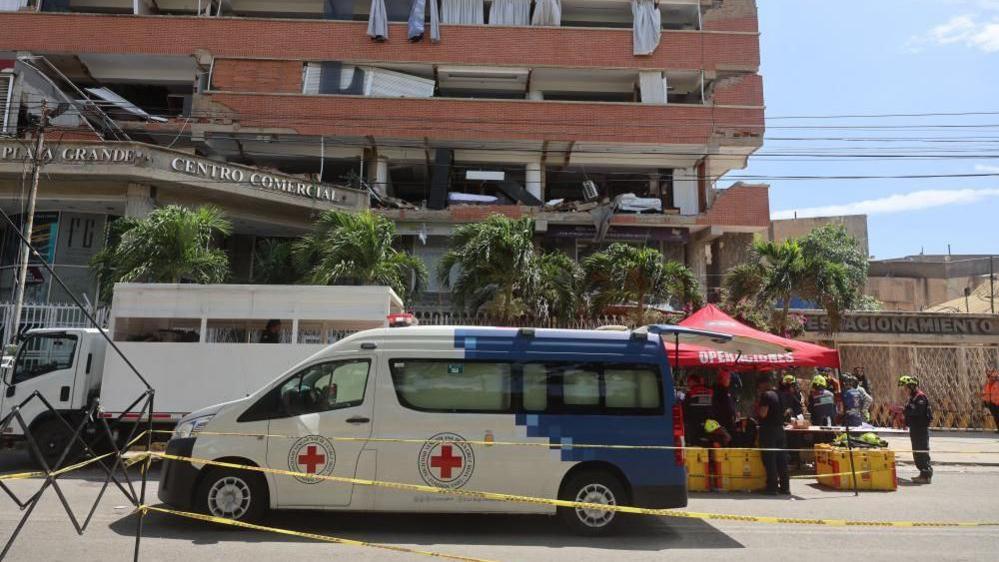 An ambulance can be seen in front of the Playa Grande shopping centre in Catia La Mar. A red tent which serves as an operation centre can also be seen. The car park is to the right of the building and only the entrance is left standing. 