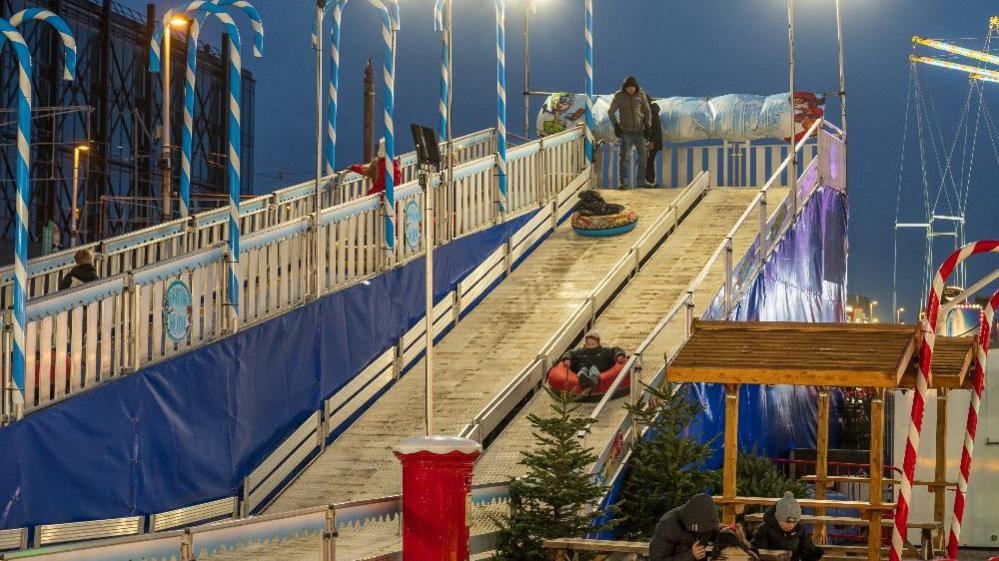 A snow slide ride decorated with blue and white candy canes on Blackpool Promenade showing youngsters going down it and other children climbing up to the top.