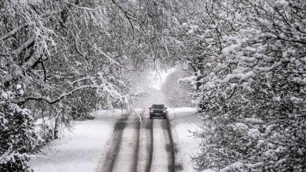 A car drives along an icy road, with snow-covered trees on either side.
