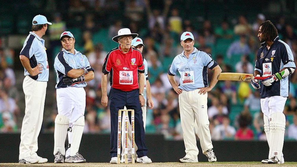 Sir Michael Parkinson - wearing a red shirt and blue trousers and a white hat - stands behind stumps, with five players surrounding him at the Sydney match in February 2009