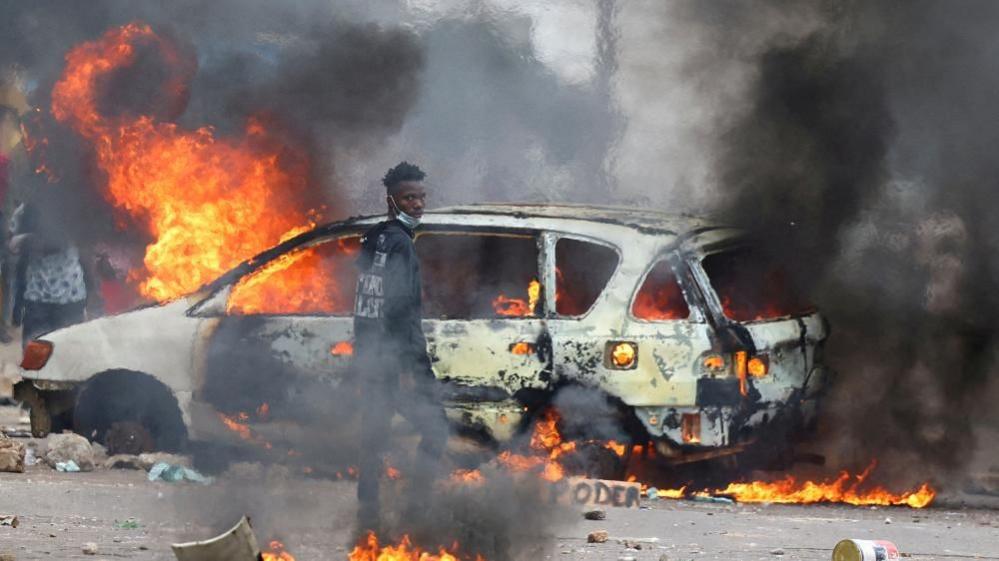 A protester stands in front of the shell of a car that is on fire. Flames and smoke billow out of the vehicle. 