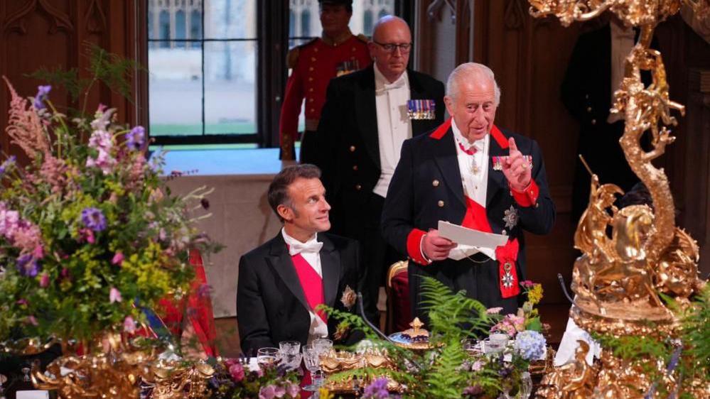 Emmanuel Macron sits at the banquet table looking up at King Charles as he gives a speech while pointing his finger.