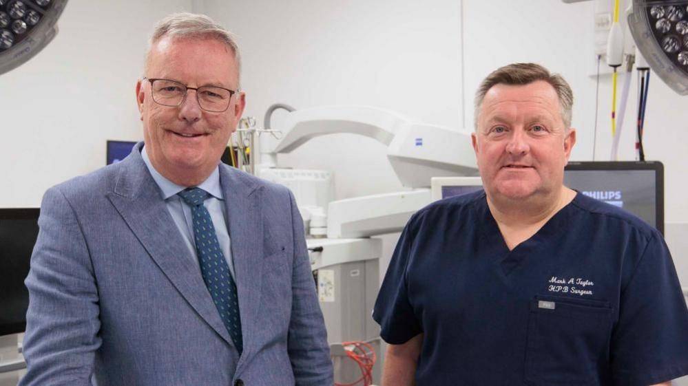 Two men stand in a surgical ward with surgical lights and machines. The man on the left is wearing a light blue suit, light blue shirt and teal tie. He has white hair, is wearing glasses and is smiling. The man on the right has brown and white hair and is wearing dark blue medical scrubs.