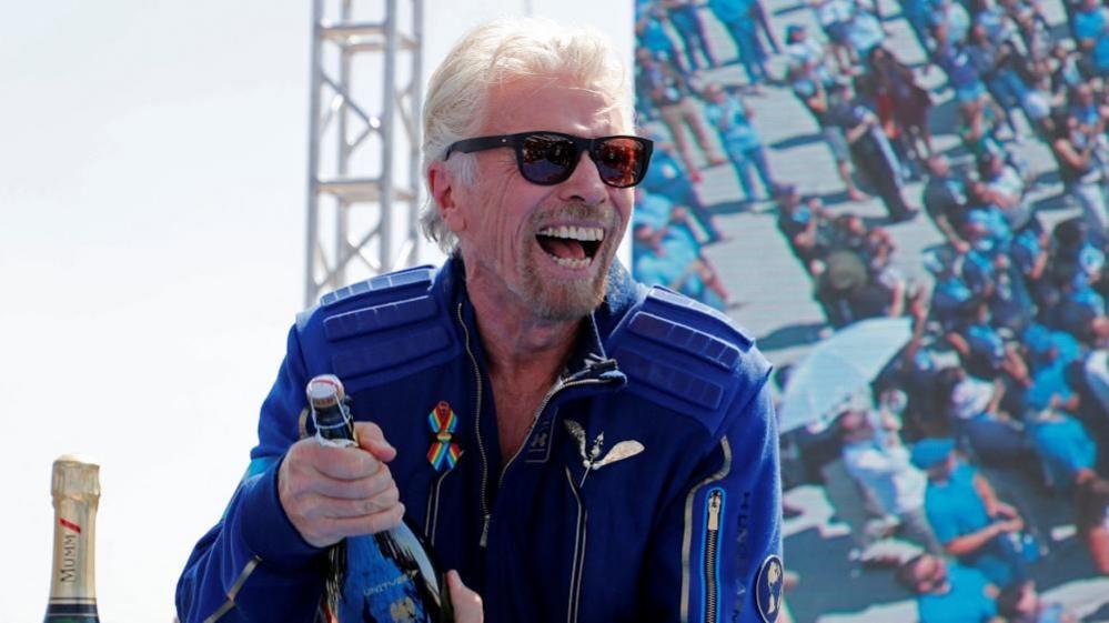 Richard Branson with medium-length white hair and dark glasses wearing a blue jacket and carrying a bottle of champagne. There are people in a grandstand behind him, some with umbrellas.