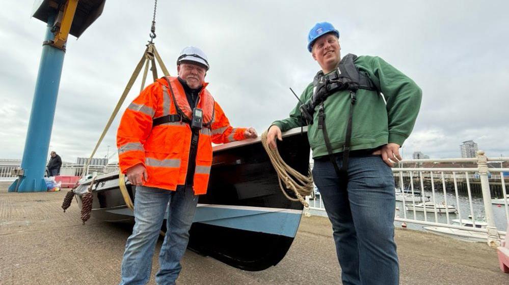 Peter Johnson and Philip Smith stand in front of the Lilian boat. It is a small open fishing boat painted black, white and blue.