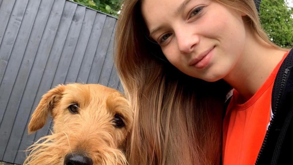 A young woman with long fair hair takes a selfie with a caramel-coloured dog. 