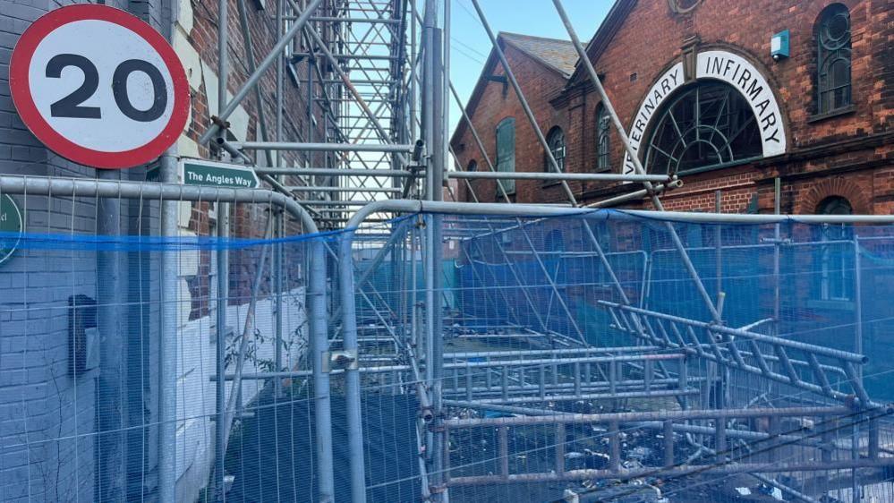 Blue netting hung over temporary fencing, surrounding a structure of scaffolding. The burned out Haven Bridge pub is on the left, with a 20mph road sign nearby, and on the right the former Shipley's Veterinary Infirmary, with the name emblazoned in an arched lintel.