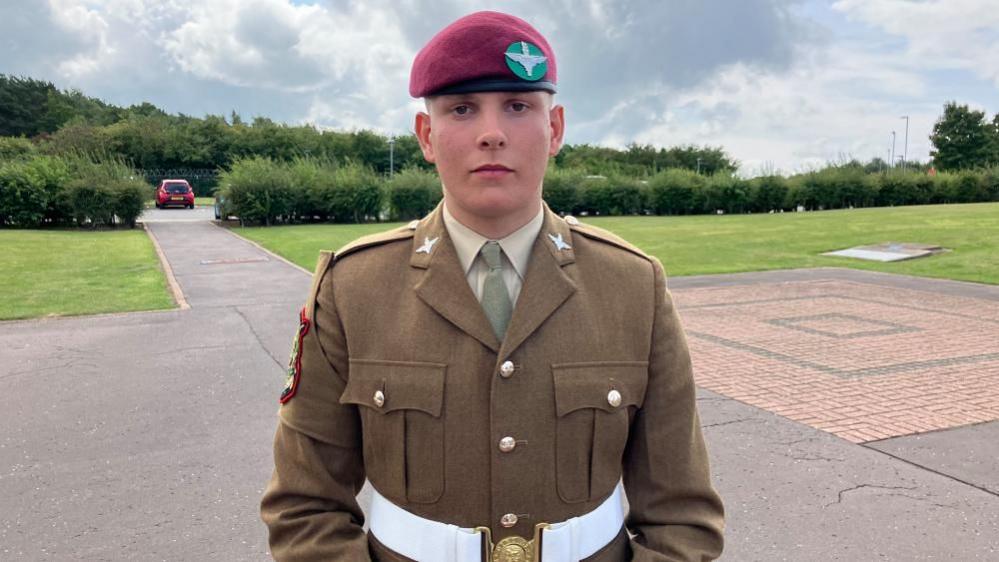 A 16 year old young man wearing formal military uniform including a red beret looks into the camera.