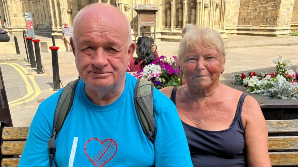 An older man with thin hair sits on a bench next to a woman with white and blond hair. York Minster is in the background.