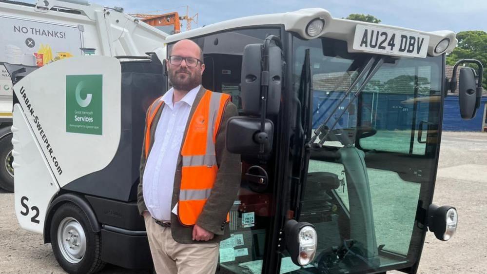 Councillor Paul Wells from Great Yarmouth Borough Council stood in front of a road sweeper vehicle. He is bald and has a beard and black wide-framed glasses. He is wearing a hi-vis jacket over the top of his suit.