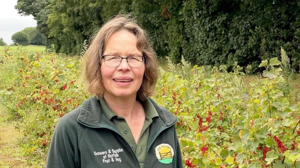 Emma Tacon stands in a field of berries wearing a green fleece, branded with her farm shop logo. Emma has long straw-blonde hair.