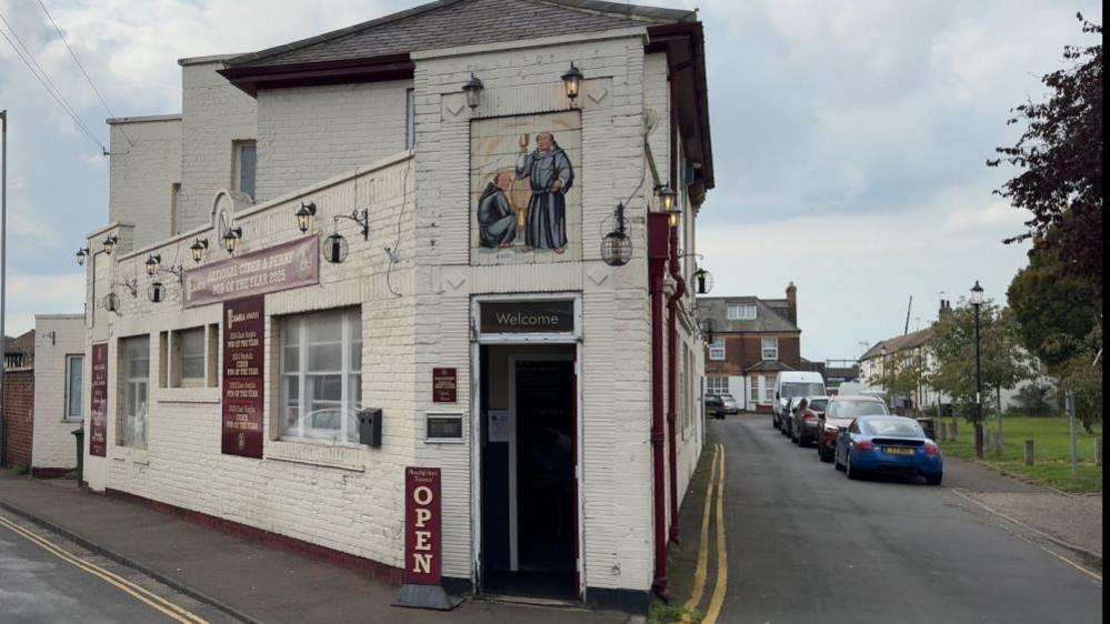 The Blackfriars Tavern is a Victorian brick-built building painted white, but modernised in the Art Deco style with features including ceramic work, one displaying a pair of monks in black habits, holding glasses of drink. To the right of the pub is a road, with carks parked near a grass are which is beside Great Yarmouth's medieval town wall.
