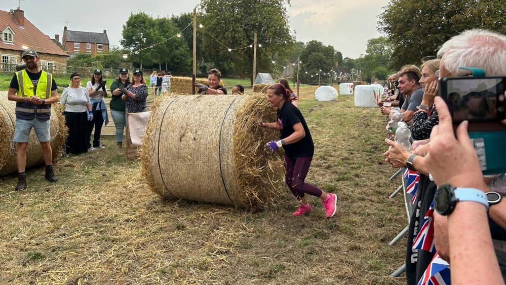 Four women wearing gloves push a large straw bale along a grass course with white static bales acting as obstacles in the background and spectators to the right and left. Some are taking photos. There is a string of lights and trees in the background.