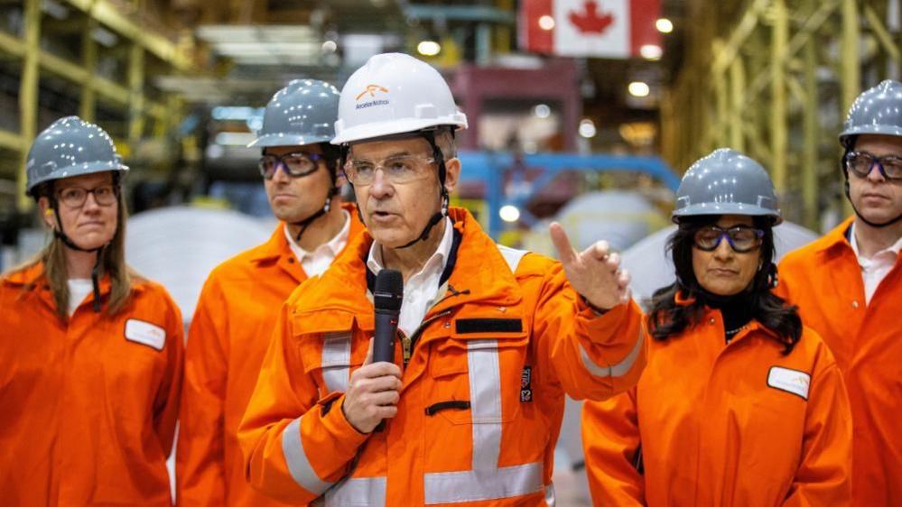 Canada's Prime Minister-designate Mark Carney speaks during his visit to the ArcelorMittal Dofasco steel mill in Hamilton, Ontario. He is speaking into a microphone and wearing a hard hat and orange jumpsuit. He is flanked by four other Liberals wearing similar outfits. 