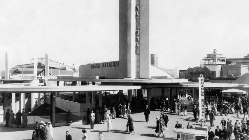 An old black and white image of people milling around the front of a tall, concrete tower with the word 'Grand National' on it. The rails of a rollercoaster can be seen behind the tower.