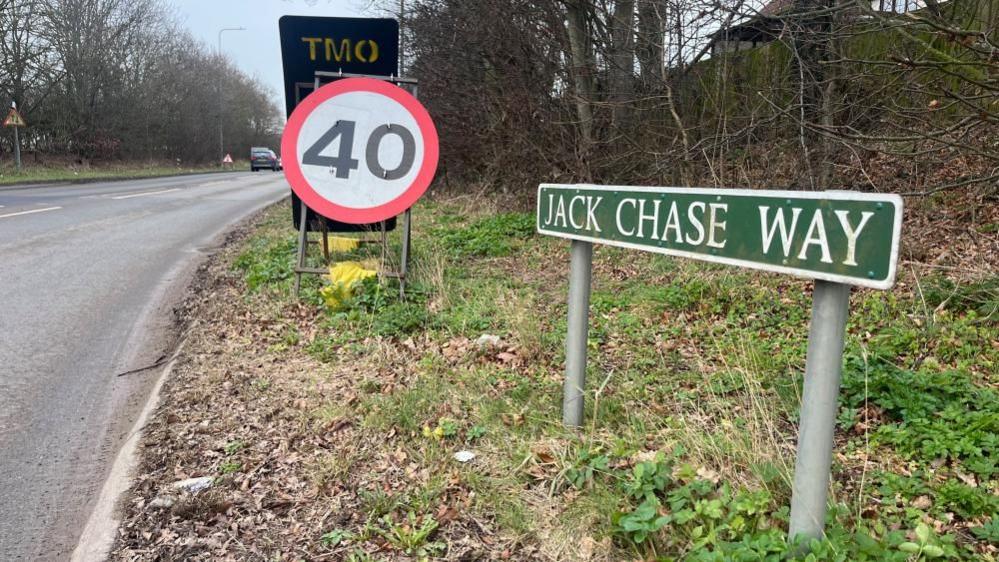 A road sign depicting Jack Chase Way with a 40mph temporary speed limit sign visible next to it. There is undergrowth on the roadside, and the road is visible to the left of the image with cars in the distance.