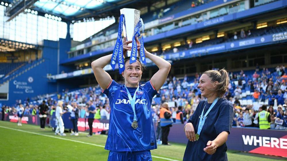 Lucy Bronze wearing a blue Chelsea strip on the pitch at Stamford Bridge wearing a medal and holding the silver WSL trophy on her head. It has blue ribbons coming off it. She's smiling and looking to the right. People can be seen in the crowd behind her.