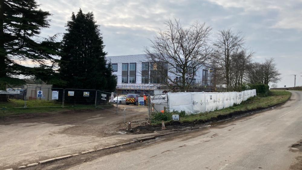 Building of a school, showing a road, trees, and bushes, then a white box building being constructed, with fencing round it, and cars parked outside it. 