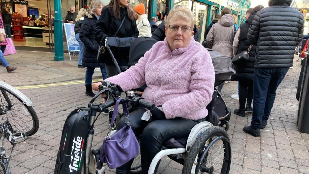 A woman with short white hair and glasses sits in a tricycle-style wheelchair wearing a pink fleece and black trousers carrying a purple handbag. 